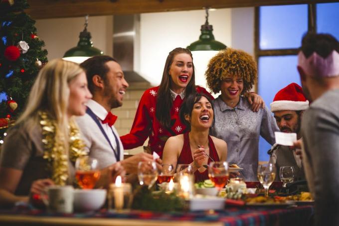 grupo de amigos rindo sentados ao redor de uma mesa comendo e bebendo no Natal um homem está lendo uma piada de uma festa de biscoitos de Natal usam chapéus e suéteres de Natal