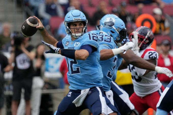 quarterback de futebol canadense com uniforme azul fazendo um passe enquanto a linha ofensiva segura um jogador defensivo em vermelho e branco