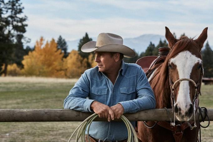 kevin costner em yellowstone ao lado de um cavalo encostado em uma cerca com corda nas mãos vestindo uma camisa jeans azul desbotada e chapéu de cowboy bege