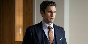 wes bentley em um terno cinza escuro de três peças com uma gravata marrom estampada e camisa listrada de branco e azul parado em um escritório com as mãos nos bolsos