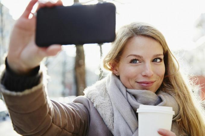 mulher bonita andando pelas ruas da cidade no inverno tirando uma selfie e segurando um café para viagem, retroiluminado