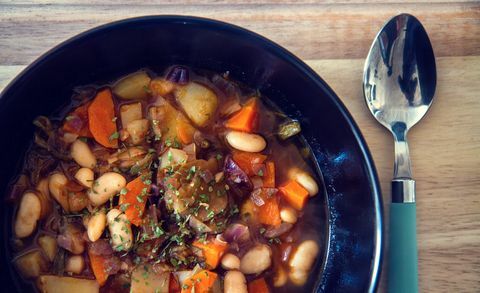 Sopa de feijão branco com cebola, cenoura, batata e legumes em um prato preto com um pouco de salsa por cima e colher ao lado.