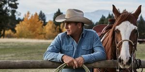 kevin costner em yellowstone ao lado de um cavalo encostado em uma cerca com corda nas mãos vestindo uma camisa jeans azul desbotada e chapéu de cowboy bege