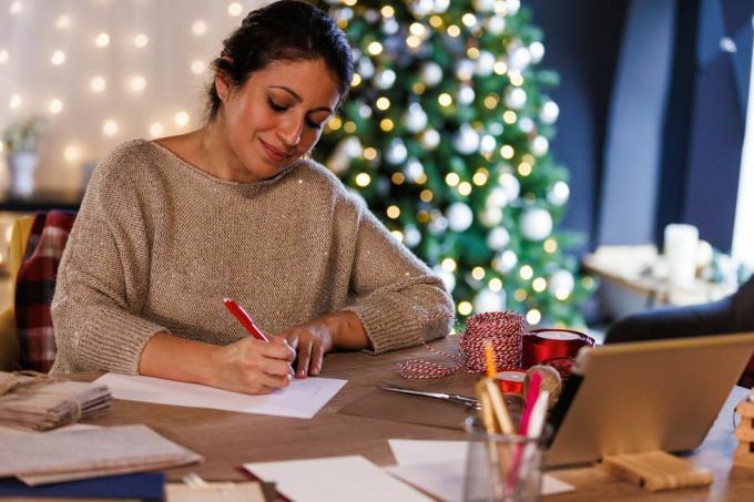 retrato de uma mulher adulta e feliz sentada à mesa, em frente a uma árvore de natal brilhante, sorrindo enquanto escreve cartões de natal e cartas para seus entes queridos