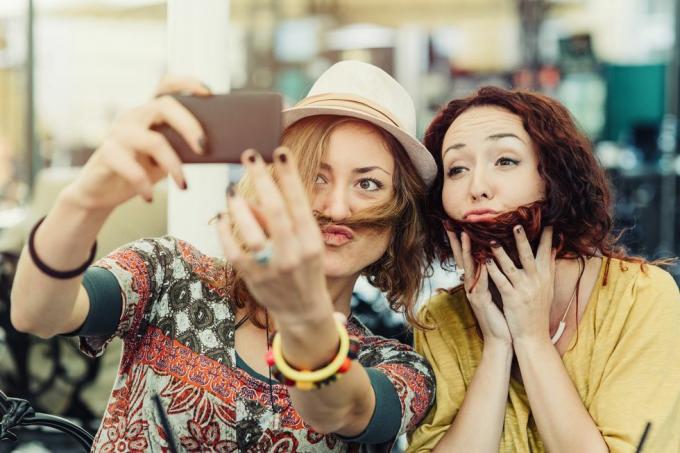 duas namoradas segurando o cabelo sobre o rosto para parecer que têm barba e bigode e tirando uma selfie