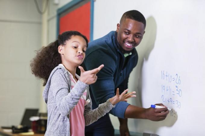 uma menina multirracial de 9 anos na aula de matemática, parada no quadro branco com seu professor, um jovem afro-americano em seu 20 anos, o professor está olhando para sua aluna do ensino fundamental, esperando enquanto ela tenta descobrir a resposta, o foco está na garota