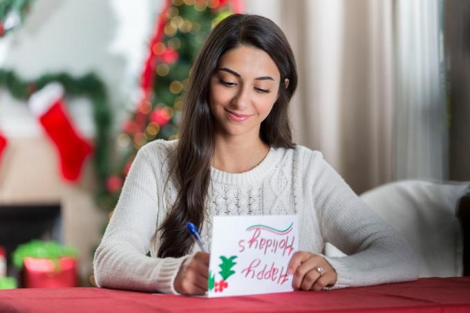 uma jovem sorridente está sentada à mesa em sua sala de estar decorada e olha para baixo, concentrada, enquanto escreve uma carta de Natal para uma amiga
